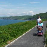 Menyw ar sgwter symudedd gyda dyn yn cerddedd ar ran darmac o lwybr arfordir cymru yn edrych allan i'r môr.