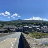 Edrych tuag at Ddolgellau o dop y Bont Fawr. Mae'r tywydd yn braf ac mae modd gweld adeiladau creigiog y dref tu hwnt i'r bont. Yn y cefndir mae mynyddoedd gwyrdd.