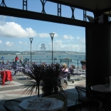 Lle eistedd awyr agored gyda’r môr y yn cefndir, a’r llun wedi’i dynnu drwy ffenest caffi.