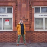 Dyn (Gareth Potter) mewn cot felen yn sefyll ar blatfform drên gydag arwydd Pontypridd.
