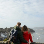 dyn a dynes yn sefyll mewn castell gyda golygfa o’r môr a chychod.