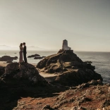 Llun priodas o gwpl yn cusanu ar ben craig ar draeth ynys Llanddwyn. Mae goleudy wen yn y cefndir.