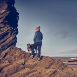 Person a chi yn sefyll ar greigiau ac yn edrych tua’r môr.