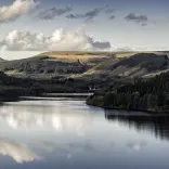 Adlewyrchiadau o awyr las gymylog ar wyneb llonydd y gronfa ddŵr, gyda choetir a mynyddoedd o’i chwmpas.