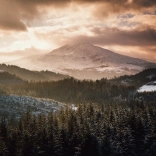 Awyr goch gymylog dros fynydd eira gyda choedwigoedd yn y blaendir.