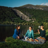 Tair menyw yn cael picnic ar lan llyn.