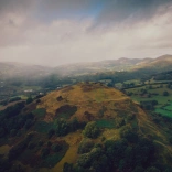 Llun o'r awyr o gastell ar fryn yng nghanol caeau gwyrdd. Mae'r awyr yn llwyd a chymylog. 