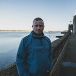Person mewn cot las yn sefyll ger dŵr a muriau hynafol tref.