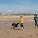 Dau oedolyn, plentyn a chi yn rhedeg o gwmpas ar draeth.