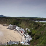 Llun o adeiladau ar y traeth, gyda bryniau gwyrdd y tu ôl. 