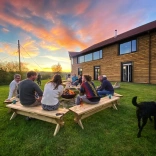 Grŵp o amgylch pwll tân y tu allan i adeilad arddull caban ar fachlud haul. 