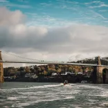 Llun o'r bont grog gyda choed a thai yn y cefndir a chwch ar Afon Menai
