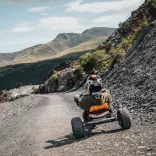 Person yn eistedd mewn go-kart yn teithio i lawr trac llechi llwyd gyda golygfeydd mynyddig yn y cefndir.
