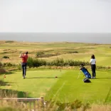 Mae dau golffwr yn sefyll ar grewn, mae'r un ar y chwith yn cymryd y swing. Gellir gweld y môr y tu hwnt i grawn y ffordd.
