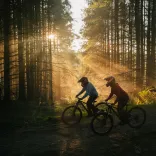 dau berson ar feiciau mynydd yn y coedwig mewn parc beiciau gyda’r haul yn disgleirio drwy’r coed.