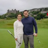 Dau o bobl yn gwenu ar gwrs golff gyda chastell yn y cefndir.