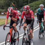 Grŵp o feicwyr ffordd proffesiynol mewn citiau rasio yn reidio'n agos gyda'i gilydd, gyda Geraint Thomas ar y blaen, ar ffordd wledig yn ystod digwyddiad beicio Tour of Britain 2025.