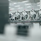 Blurred view of a factory production line with rows of identical industrial machines or workstations arranged in the background. The image is monochrome and softly focused, creating a clean, modern manufacturing atmosphere.