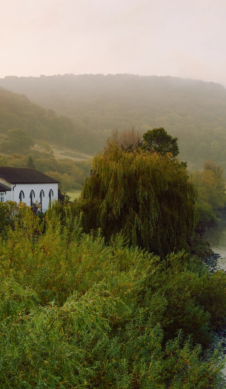 Adeilad gwyn ar lan yr afon, gyda bryniau yn y cefndir