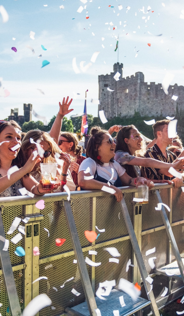 Cynulleidfa gig cerddoriaeth yn Tafwyl yn sefyll tu nôl i wahanfur gyda chonffeti o'u cwmpas a Chastell Caerdydd yn y cefndir