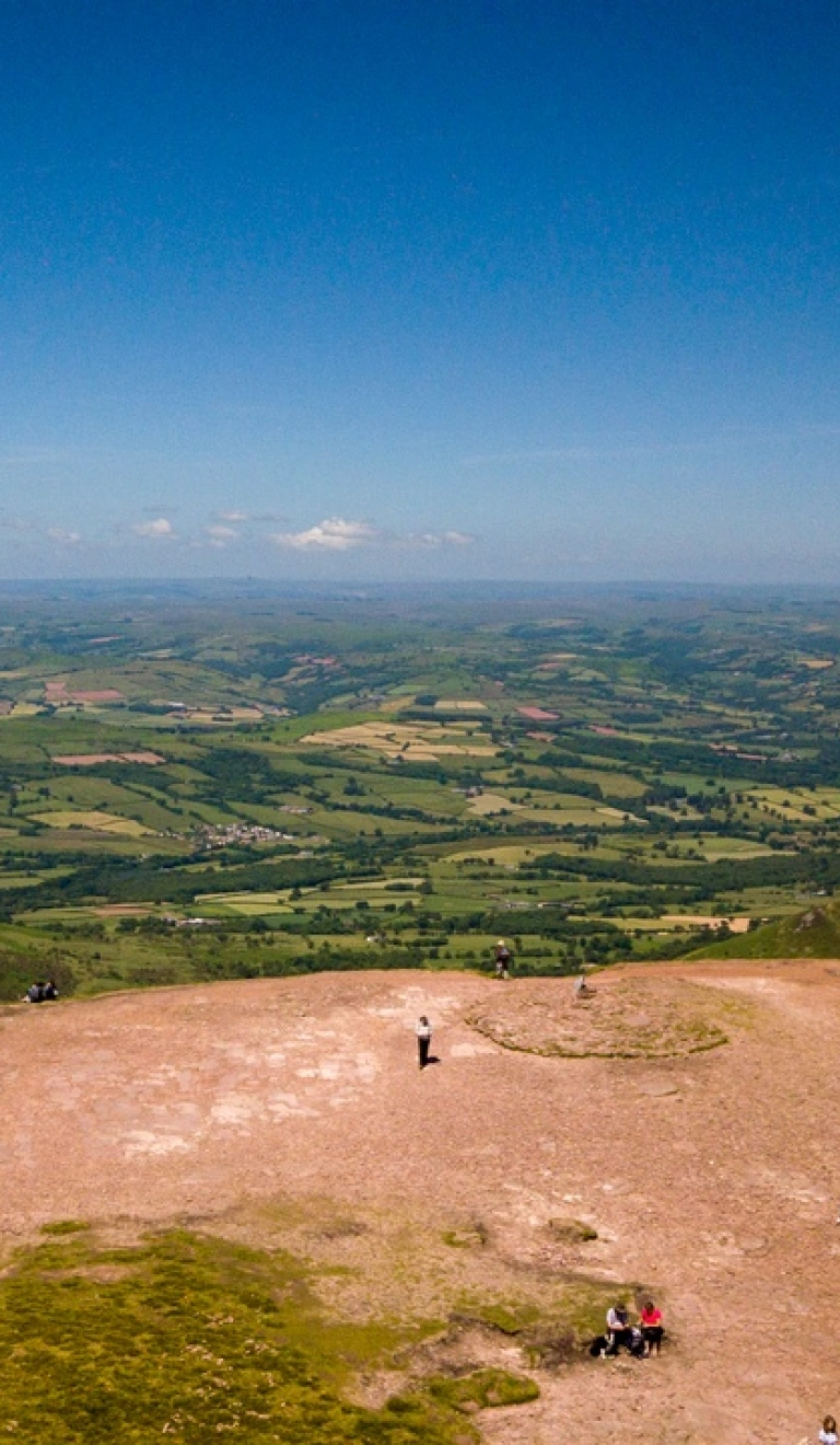 Golygfa o'r awyr o'r gwastadeddau gwyrdd islaw o ben Pen y Fan gydag awyr las.