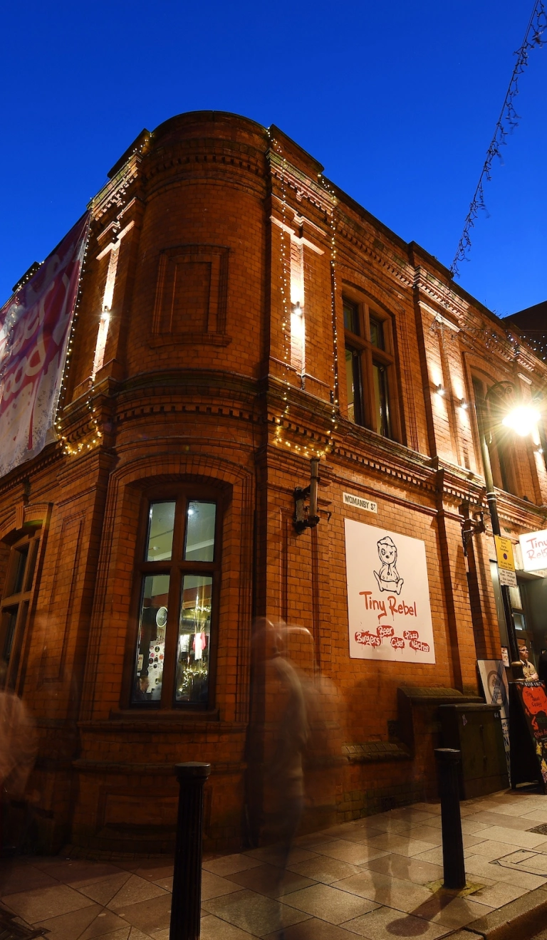 Llun o'r tu allan i Tiny Rebel yn y nos, sy'n adeilad brics coch ar gornel stryd