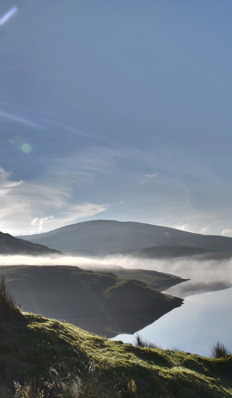 Rhywun yn edrych dros y mynyddoedd a llyn yn gynnar yn y bore.