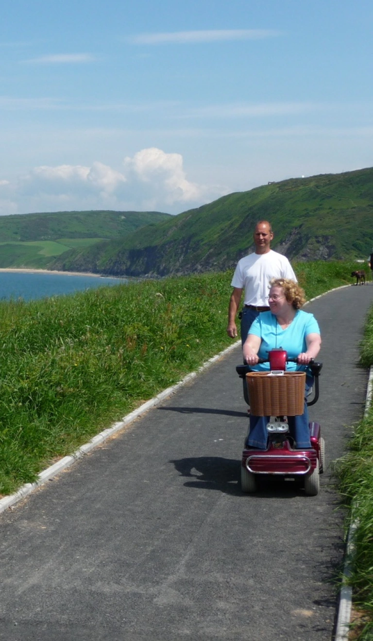 Menyw ar sgwter symudedd gyda dyn yn cerddedd ar ran darmac o lwybr arfordir cymru yn edrych allan i'r môr.