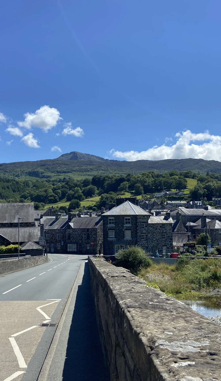 Edrych tuag at Ddolgellau o dop y Bont Fawr. Mae'r tywydd yn braf ac mae modd gweld adeiladau creigiog y dref tu hwnt i'r bont. Yn y cefndir mae mynyddoedd gwyrdd.