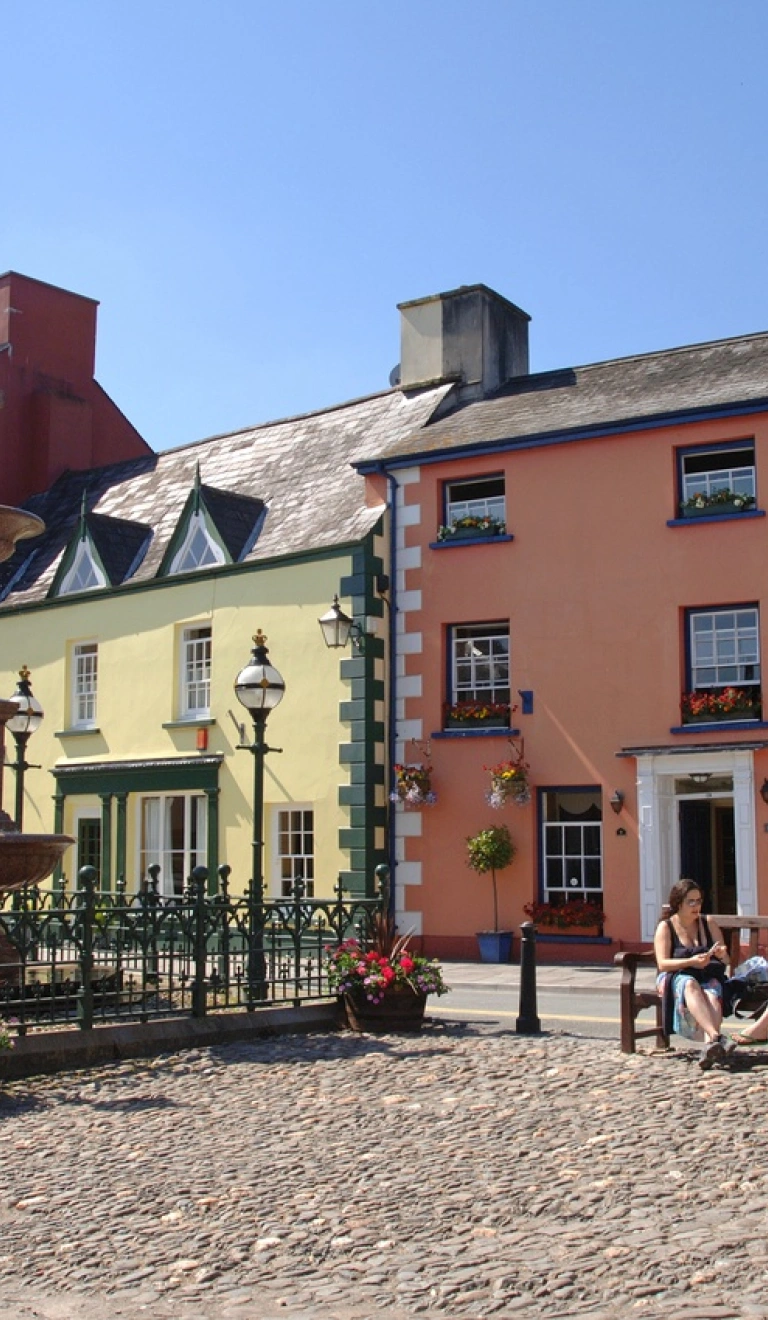Sgwâr y dref, Llanymddyfri. Mae tai mawr crand oren a melyn ar y sgwrs, a dwy ddynes yn eistedd ar fainc.