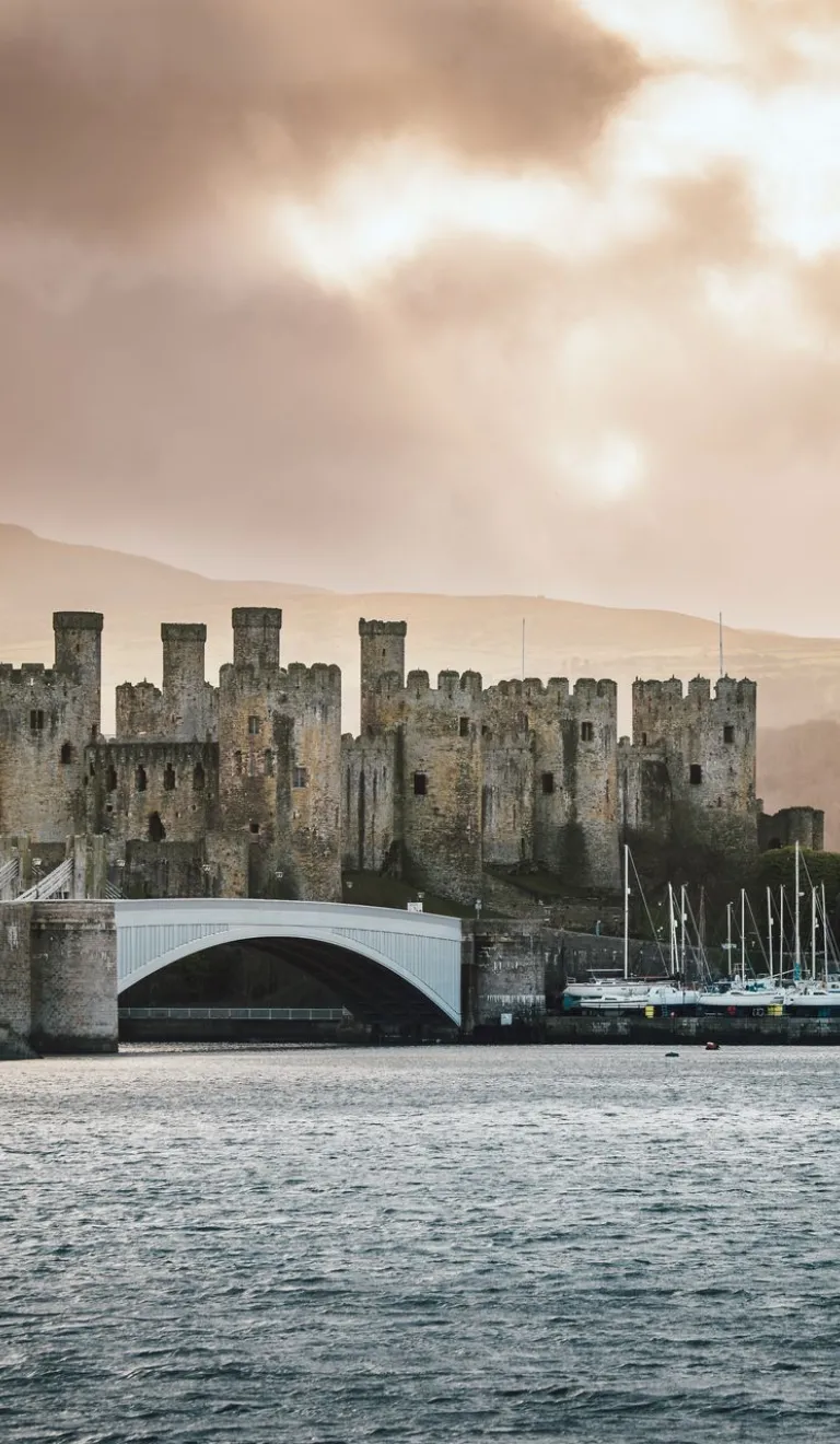 Castell anferth gyda thyrau a muriau enfawr ger marina, gydag awyr gymylog uwchben.