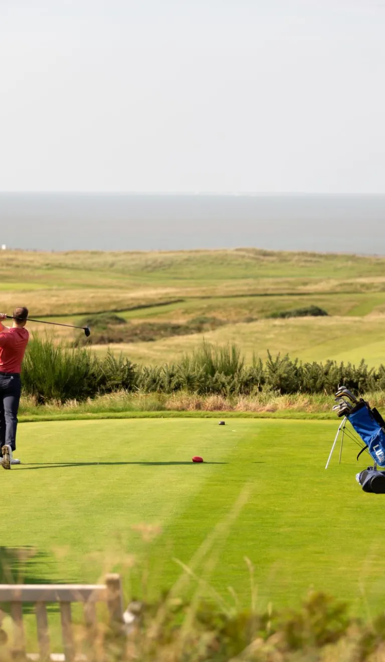 Mae dau golffwr yn sefyll ar grewn, mae'r un ar y chwith yn cymryd y swing. Gellir gweld y môr y tu hwnt i grawn y ffordd.