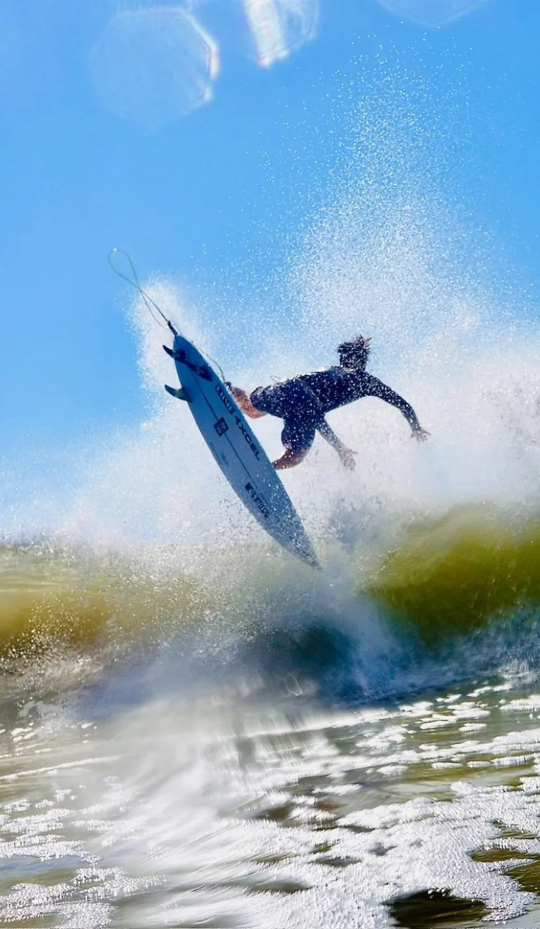 Syrffiwr yn yr awyr uwch ton sy’n torri, ac ewyn o’i gwmpas, o dan awyr las glir.